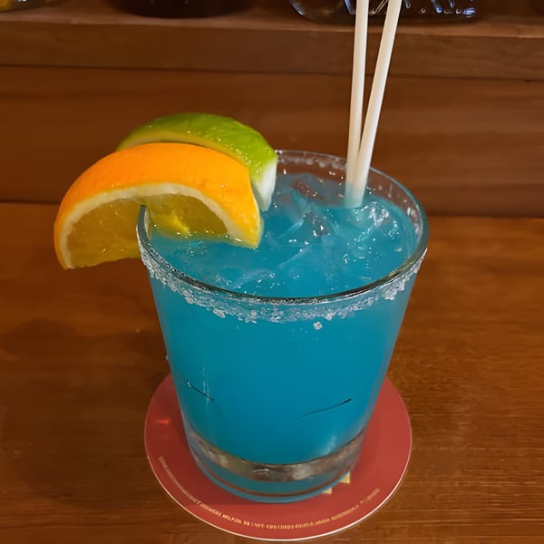 A vibrant blue cocktail with ice, garnished with orange and lime slices, and two straws.