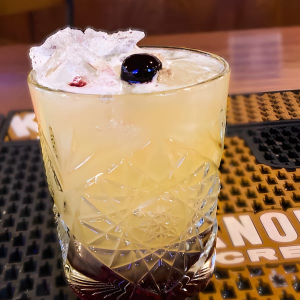 A cocktail in a cut glass, garnished with crushed ice and a dark cherry, on a bar mat.