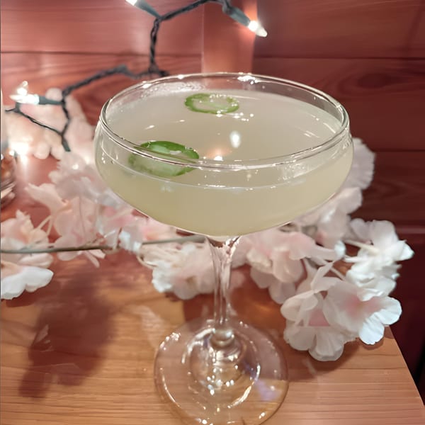 A light yellow cocktail with jalapeño slices sits on a wooden table with white flowers and string lights.