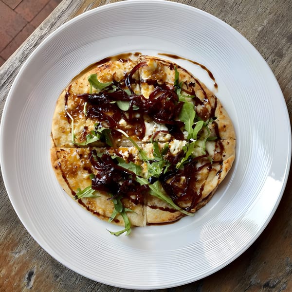 Flatbread with caramelized onions, arugula, cheese, and balsamic glaze on a white plate.
