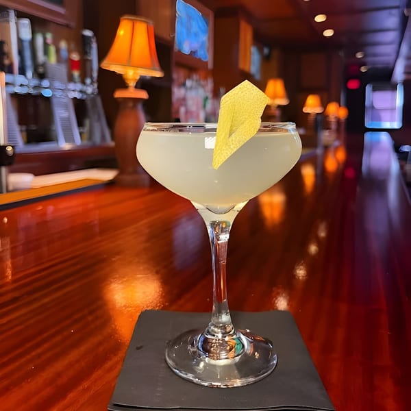 A pale cocktail with a lemon peel garnish sits on a dark bar with warm lighting.