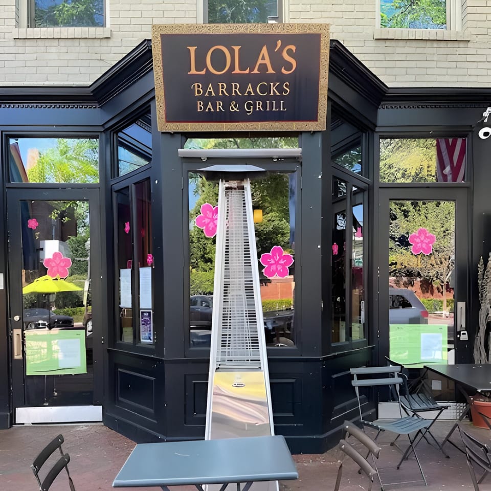 Lola's Barracks Bar & Grill storefront with outdoor seating, a patio heater, and pink flower decals.