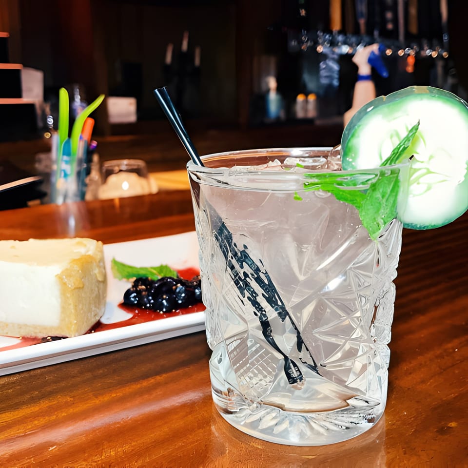 A cocktail with cucumber and mint, next to a slice of cheesecake with berries on a wooden bar.