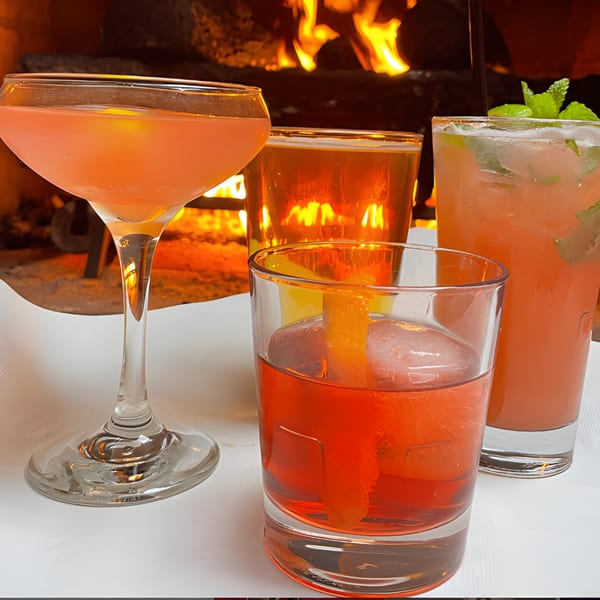 Four colorful cocktails, including a martini and an Old Fashioned, in front of a cozy fireplace.