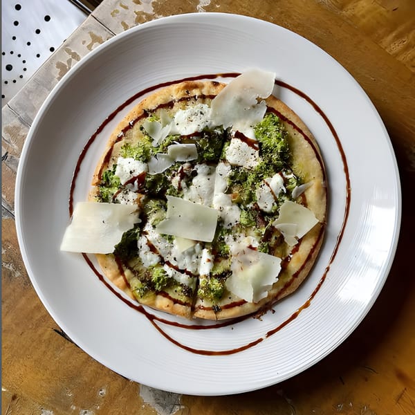 A round flatbread pizza topped with green vegetables, white cheese, and balsamic glaze on a white plate.
