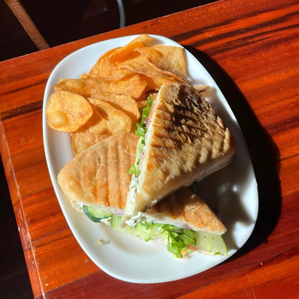 A grilled panini sandwich with chips on a white plate, on a wooden table at Lola's.