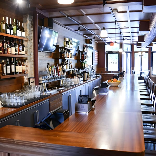 A long, polished wooden bar with bottles, beer taps, glasses, and bar stools at Lola's.